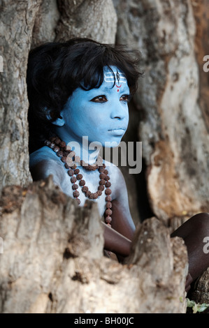 Indian boy, face painted as the Hindu god Shiva sitting in an old tree stump. Andhra Pradesh, India