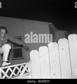 A fashionable young man jumps over a fence next to a house in a suburban neighborhood during the late 1950s. Stock Photo