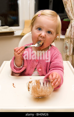 Vertical close up portrait of a baby girl getting in a mess eating chocolate ice cream in her high chair. Stock Photo