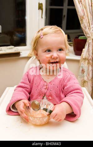 Vertical close up portrait of a baby girl getting in a mess eating chocolate ice cream in her high chair. Stock Photo