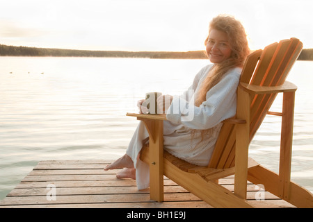 Woman in robe seated on deck chair on dock, Clear Lake, Manitoba, Canada Stock Photo