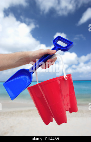 A man's hand holding a toy bucket and spade in the air on a deserted tropical beach