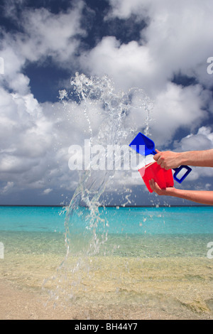 A man's hands throwing a child's toy bucket and spade full of water in the air on a deserted tropical beach Stock Photo