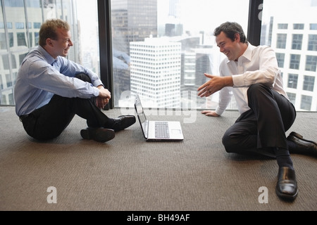 Two businessmen sitting on the office floor with a laptop computer having a discussion, smiling Stock Photo