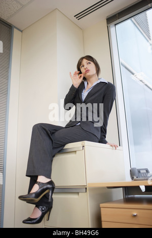 A young businesswoman sitting on top of a filing cabinet chewing gum Stock Photo