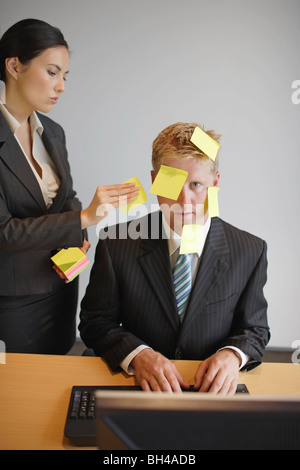 A businesswoman placing sticky notes of paper on to a businessman's face as he sits at a desk working on a computer in an office Stock Photo