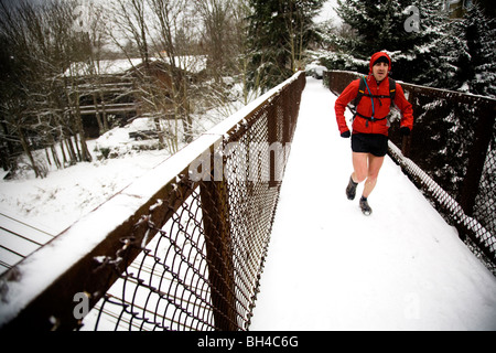 A male runner with a backpack runs across a snow-covered bridge while wearing shorts in the winter. Stock Photo