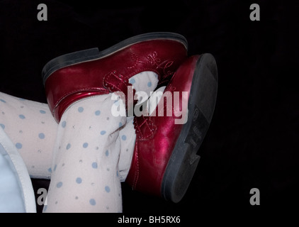 Photo shows tiny baby's feet in red shoes and polka dot tights crossed at the ankles against a black background. Stock Photo