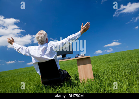 Business concept shot showing an older male executive arms raised using a computer in a green field with a blue sky & clouds Stock Photo