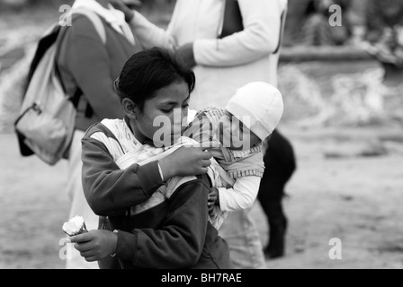 Ecuador, Otavalo, young teenage girl carrying her baby brother or sister on her back and sharing a cone of ice cream with him Stock Photo