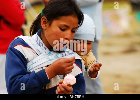 Ecuador, Otavalo, young teenage girl carrying her baby brother or sister on her back and sharing a cone of ice cream with him Stock Photo