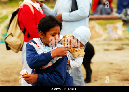 Ecuador, Otavalo, young teenage girl carrying her baby brother or sister on her back and sharing a cone of ice cream with him Stock Photo