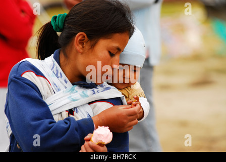 Ecuador, Otavalo, young teenage girl carrying her baby brother or sister on her back and sharing a cone of ice cream with him Stock Photo