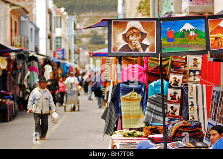 Ecuador, Otavalo, view of a series of realistic paintings exhibited at a stall on a street market with people walking in market Stock Photo