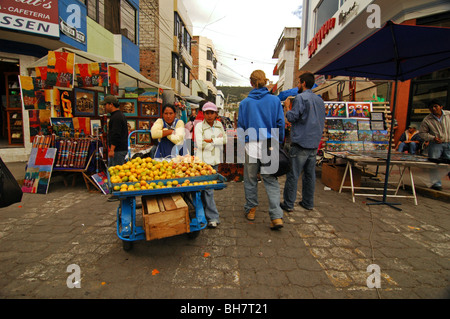 Ecuador, Otavalo, rear view of a couple of tourists passing by two young girls selling fruits on their blue cart at the weekly m Stock Photo