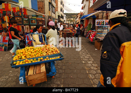 Ecuador; Otavalo; rear view of a couple of tourists passing by two young girls selling fruits on their blue cart at the weekly m Stock Photo