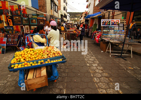 Ecuador, Otavalo, rear view of a couple of tourists passing by two young girls selling fruits on their blue cart at the weekly m Stock Photo