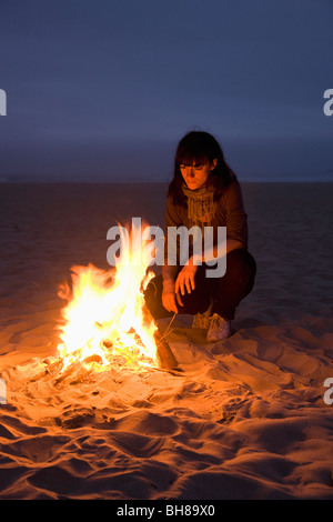 A woman staring into a fire burning on a beach Stock Photo