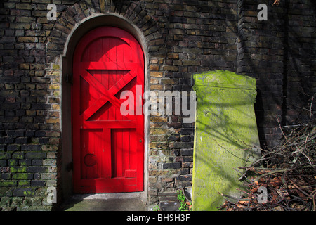 Bright red door in an old church yard wall with a large green algae covered gravestone next to the door, lengthy inscription Stock Photo