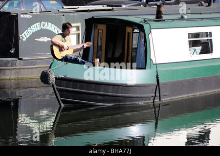 Young man playing a guitar on a canal boat on the river Lea, Hertford, Hertfordshire, England, UK. Stock Photo