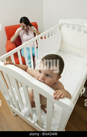 Young woman on arm chair holding her head and baby boy in crib Stock Photo