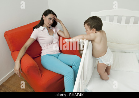 Young woman on arm chair holding her head and baby boy in crib Stock Photo