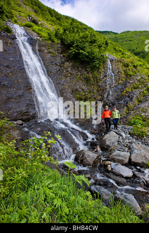 Couple walking along the shoreline in front of a waterfall, Shoup Bay State Marine Park, Prince William Sound, Alaska Stock Photo