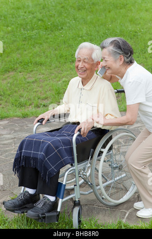 Japan, Tokyo Prefecture, Senior man sitting on wheelchair, woman using mobile phone, smiling Stock Photo