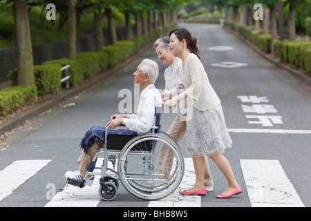 Japan, Tokyo Prefecture, Woman pushing senior man in wheelchair, crossing road, side view Stock Photo