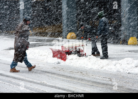 Two men shoveling snow with an automatic plow while man is walking crossing the street during heavy snow fall in bad weather bli Stock Photo