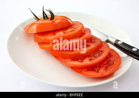 One red, ripe, juicy beef tomato cut into six slices, with knife on white plate with white background. Stock Photo