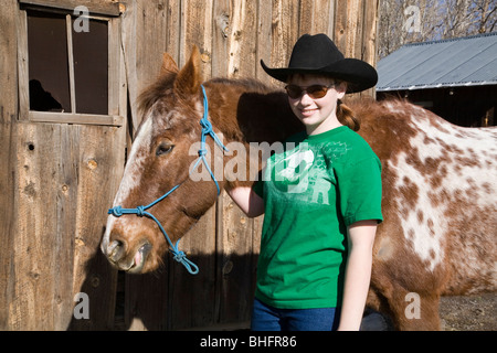 A young cowgirl wearing a ten gallon hat and her roan appaloosa horse in front of an old barn. Stock Photo