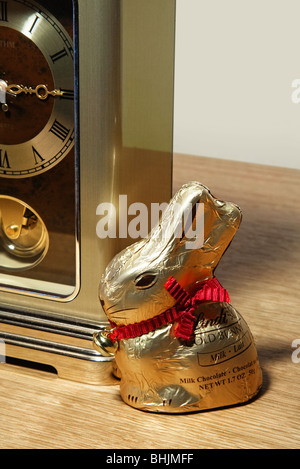 A Lindt gold foil wrapped chocolate Easter rabbit sitting next to a clock. Stock Photo