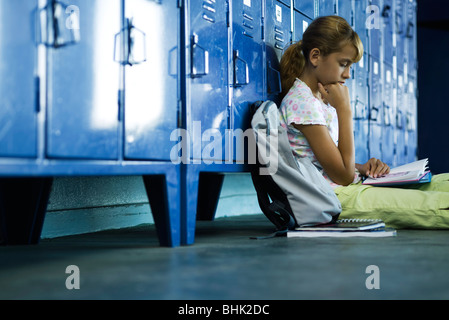 Female junior high student sitting on hall floor leaning against lockers reading book Stock Photo