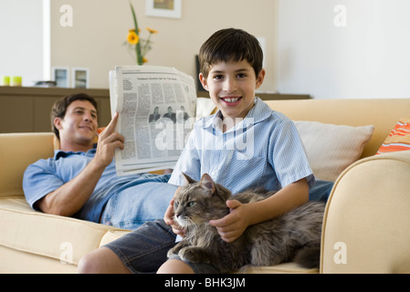 Young boy with pet cat on lap, father reading newspaper in background Stock Photo