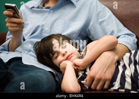 Father text messaging while son naps with head in his lap Stock Photo