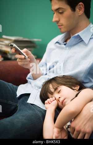 Young man text messaging while son naps with head in his lap Stock Photo