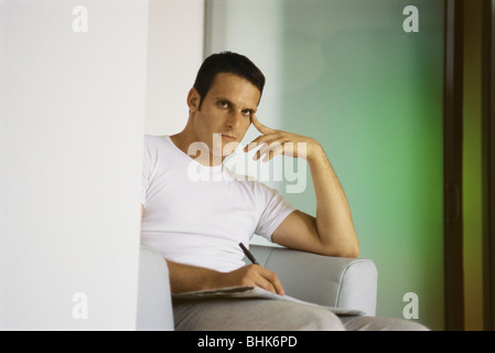 Young man sitting in armchair, holding pen and newspaper, looking at camera Stock Photo