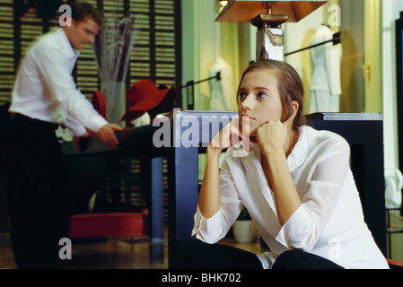 Young woman sitting with hands under chin in clothing store, man looking at table in background Stock Photo