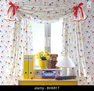 Close-up of floral curtains on window above desk with yellow box-files and toy typewriter in playroom with floral wallpaper Stock Photo