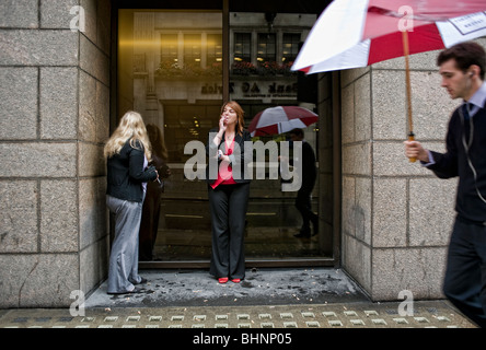 A female office worker shelters from the rain in a doorway to smoke a cigarette outside her place of work, London. Stock Photo