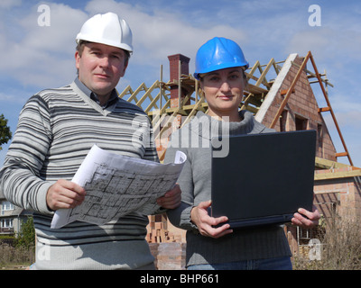 Female and male building engineers standing against unfinished brick house, man holding building plans and woman holding laptop Stock Photo