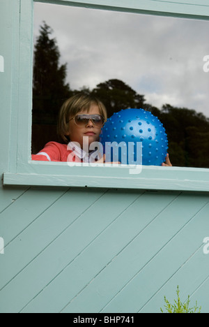 a small boy with fashionable adult sunglasses and holding onto a metallic blue football gazes out of a window longing to play Stock Photo