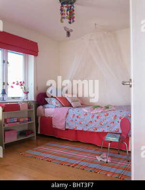 Pink blind on window in child's bedroom with coronet and white voile drapes above bed with blue floral bedlinen and pink valence Stock Photo