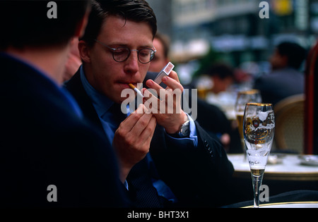 Striking a light in an outdoor café, a young businessman puts a match to his cigarette as a colleague talks in Frankfurt. Stock Photo