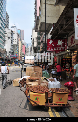 Busy street scene, shops, shoppers, flat carts, boxes, bamboo drying trays of cashew kernels, Des Voeux Road West, Hong Kong Stock Photo