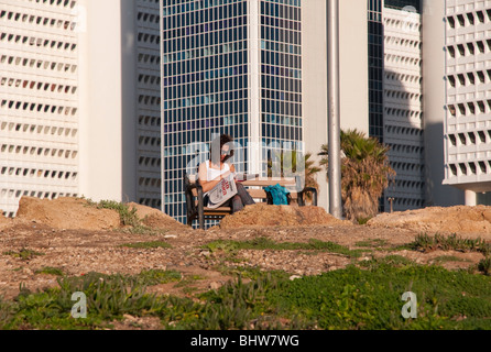 woman reading newspaper on sea front promenade in tel aviv with textile complex in bkgd Stock Photo