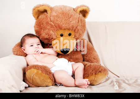 Cute Caucasian Hispanic unisex baby in arms of a big brown stuffed teddy bear sitting on couch. Stock Photo