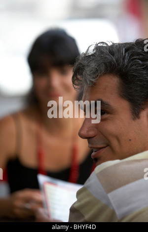 early thirties hispanic man smiling sitting with partner at outdoor cafe table looking for waiter service Stock Photo