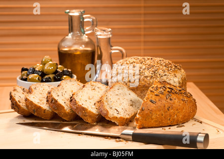Freshly cut and sliced rustic seeded bread with a knife and a bowl of green & black olives, olive oil & balsamic vinegar Stock Photo
